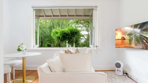 Una habitació amb un sofà blanc, una taula rodona amb un ram de flors, una finestra amb vista a vegetació i un televisor penjat a la paret que mostra una escena de ponent.