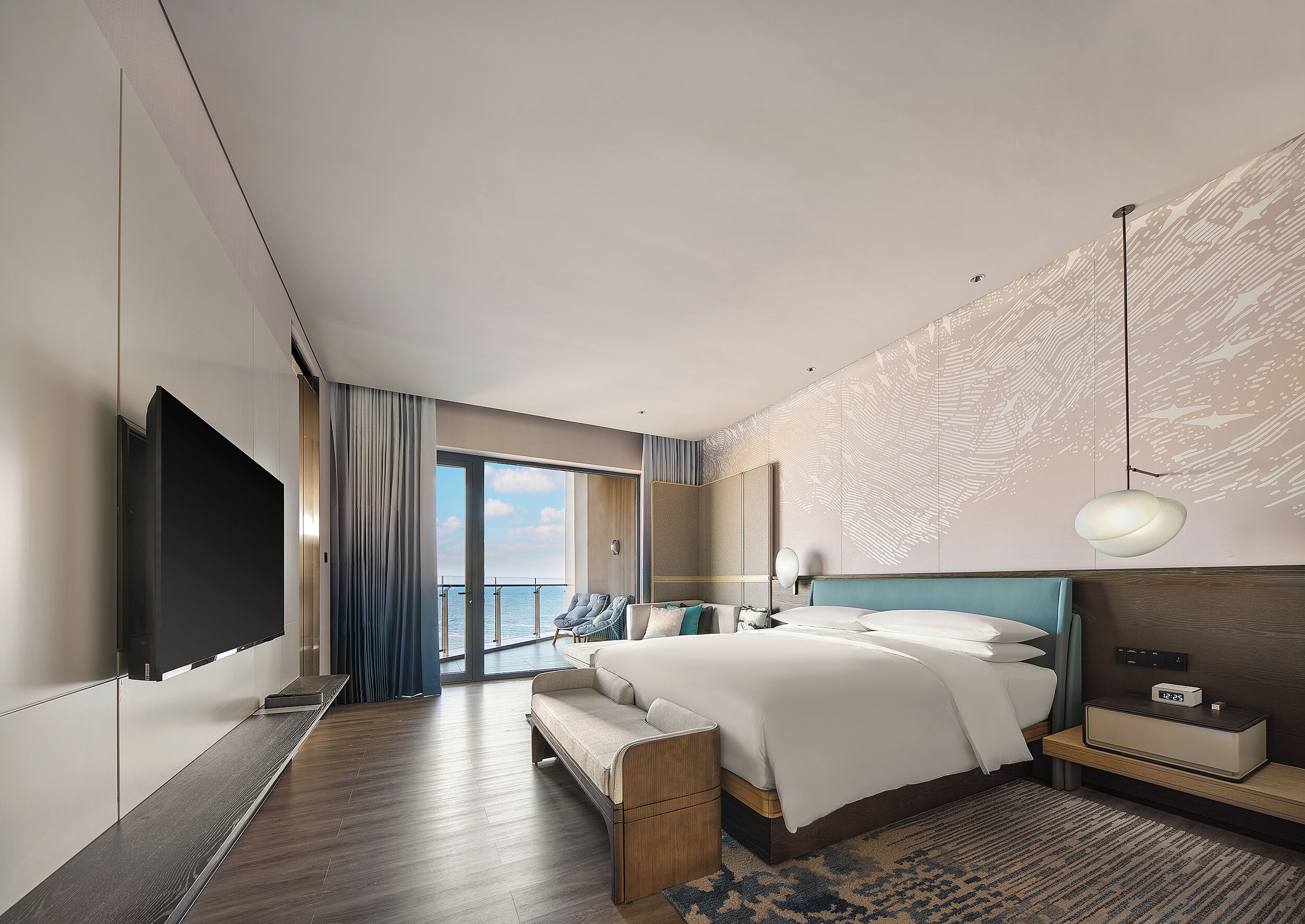 A modern hotel room with a large bed, a flat-screen TV, a seating area with a view of the sea, and a patterned wall.
