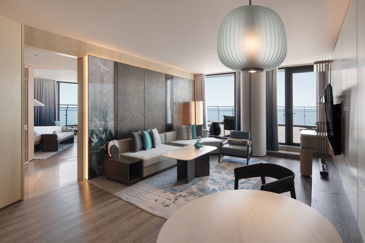 A modern living room with a large sofa, a coffee table, and a dining table. There is a large window with a view of the sea.