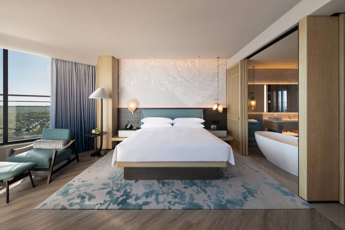 A modern hotel room with a large bed, a sitting area with a chair, a desk, and a view of the city.