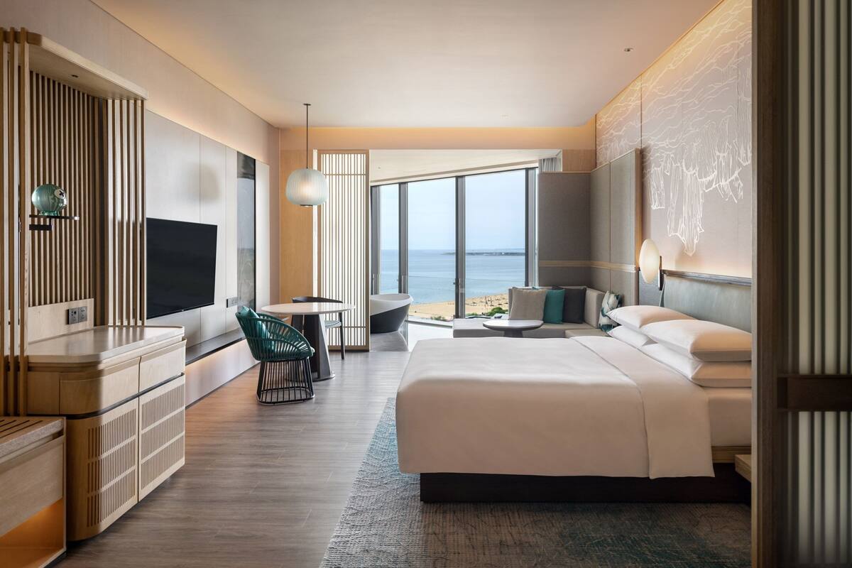A modern hotel room with a large bed, a desk, a chair, and a view of the beach.
