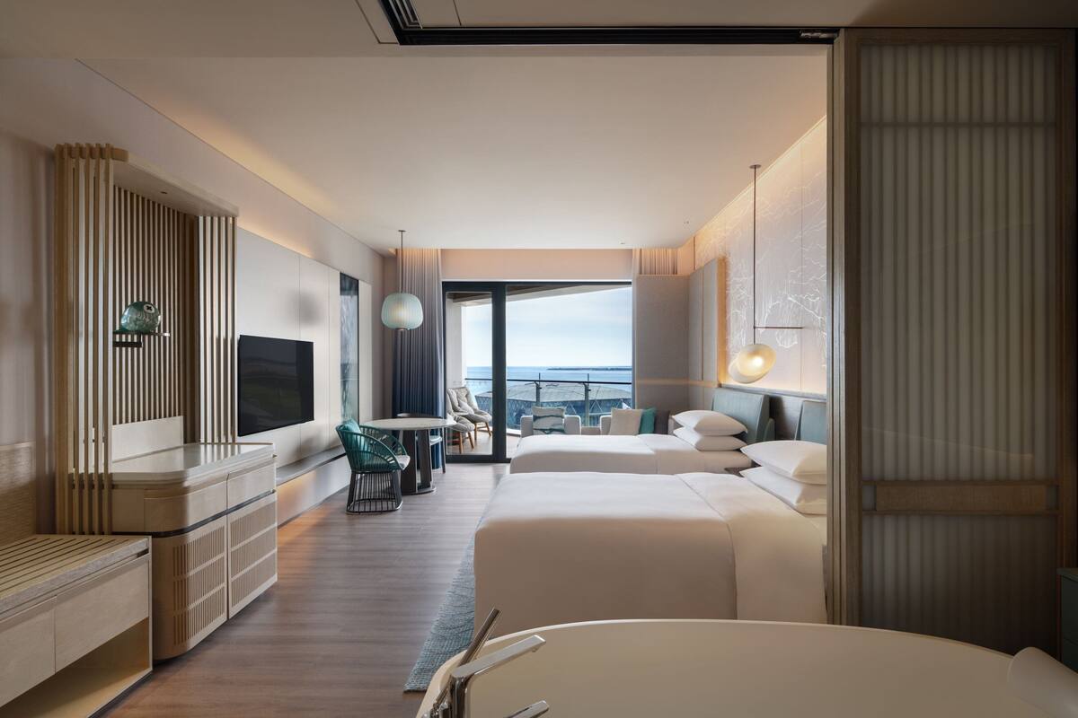 A modern hotel room with a large bed, a desk with a chair, a TV, and a view of the ocean.