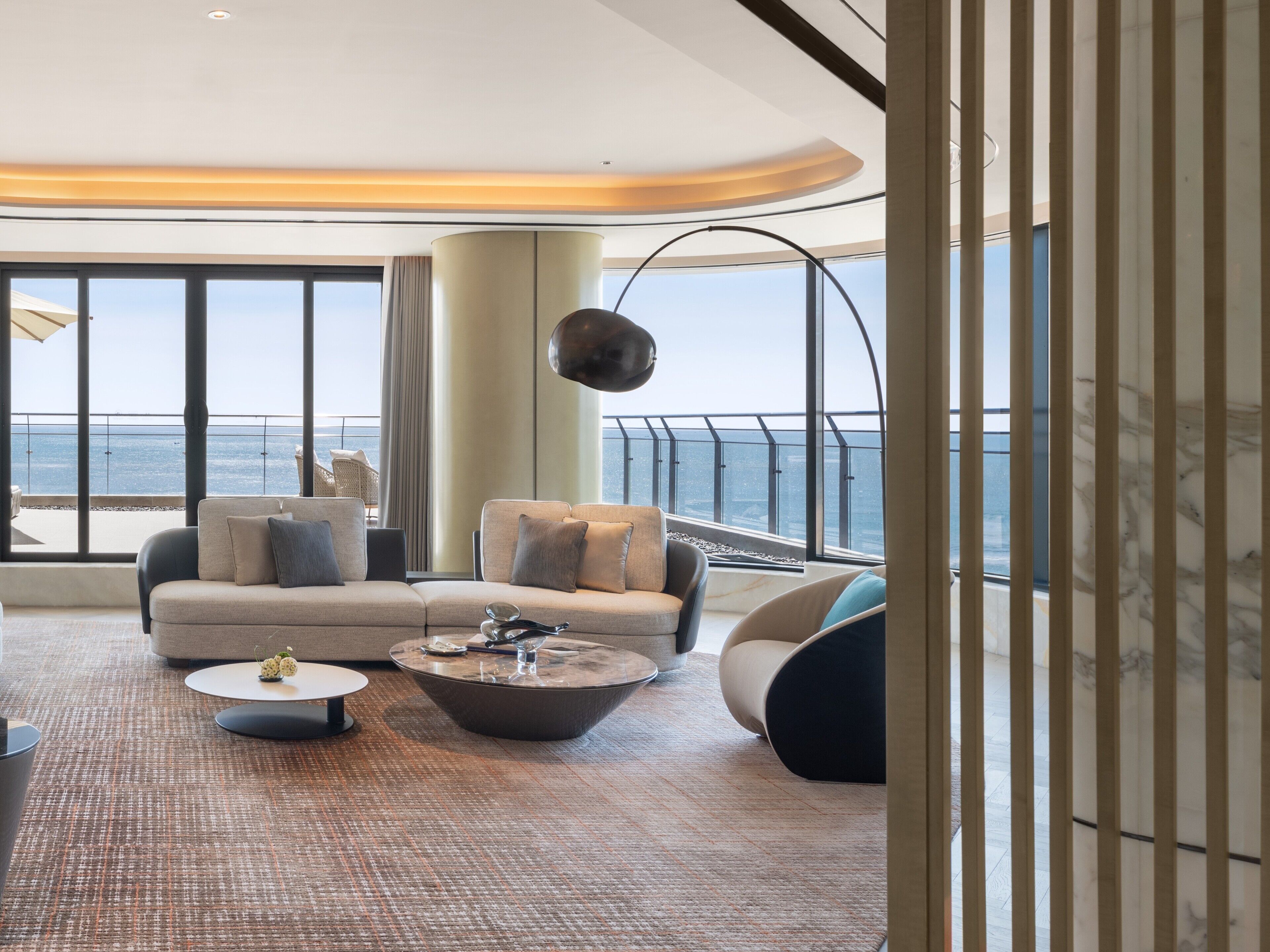 A modern living room with a sofa, armchairs, a coffee table, and a large window offering a view of the sea.