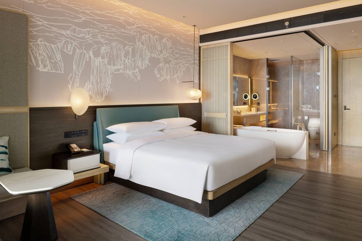 A modern hotel room with a large bed, bedside table, and a bathroom with a bathtub and shower.