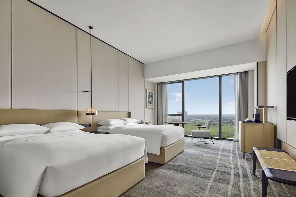 Ein Hotelzimmer mit zwei Betten, einem großen Fenster mit Blick auf die Stadt, einem kleinen Tisch und einem Sessel.