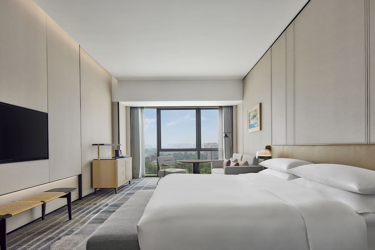 Ein modernes Hotelzimmer mit einem großen Bett, einem Flachbildfernseher, einem kleinen Tisch und Blick auf die Stadt durch große Fenster.