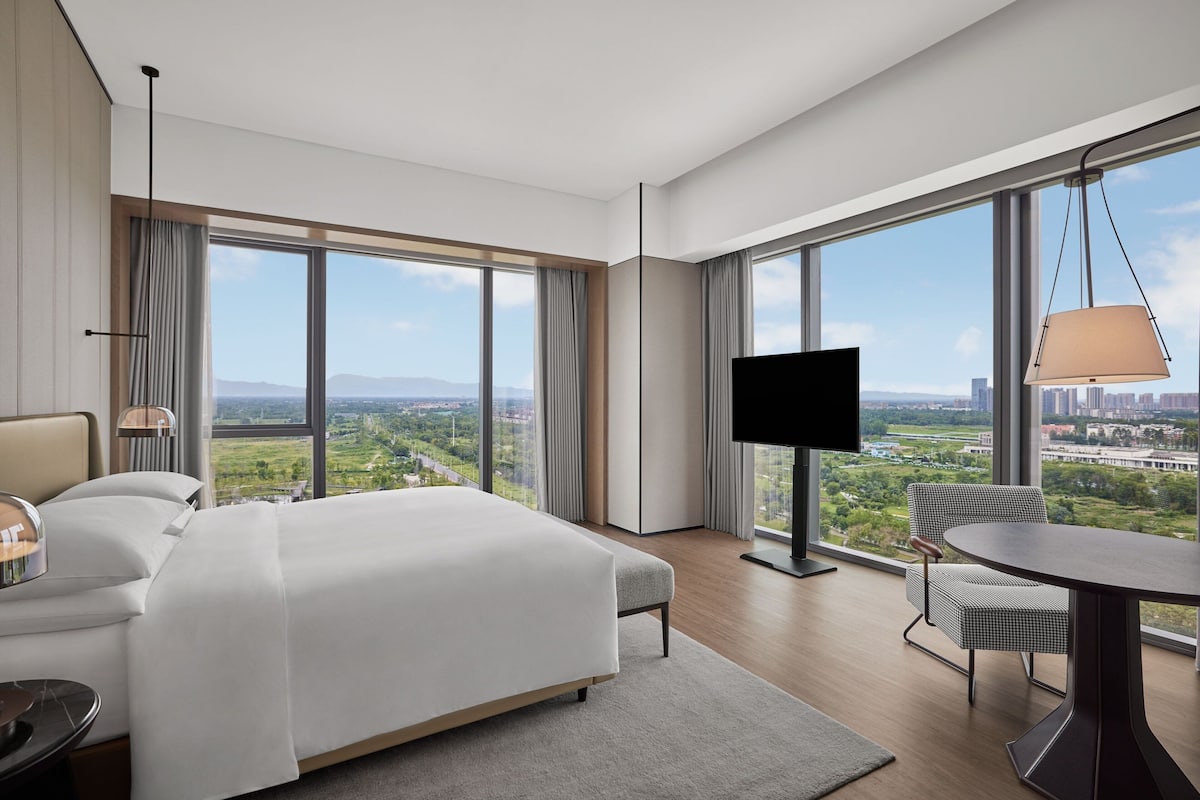 Ein modernes Hotelzimmer mit einem großen Bett, einem Fernseher, einem Esstisch und Blick auf Stadt und Grünflächen.