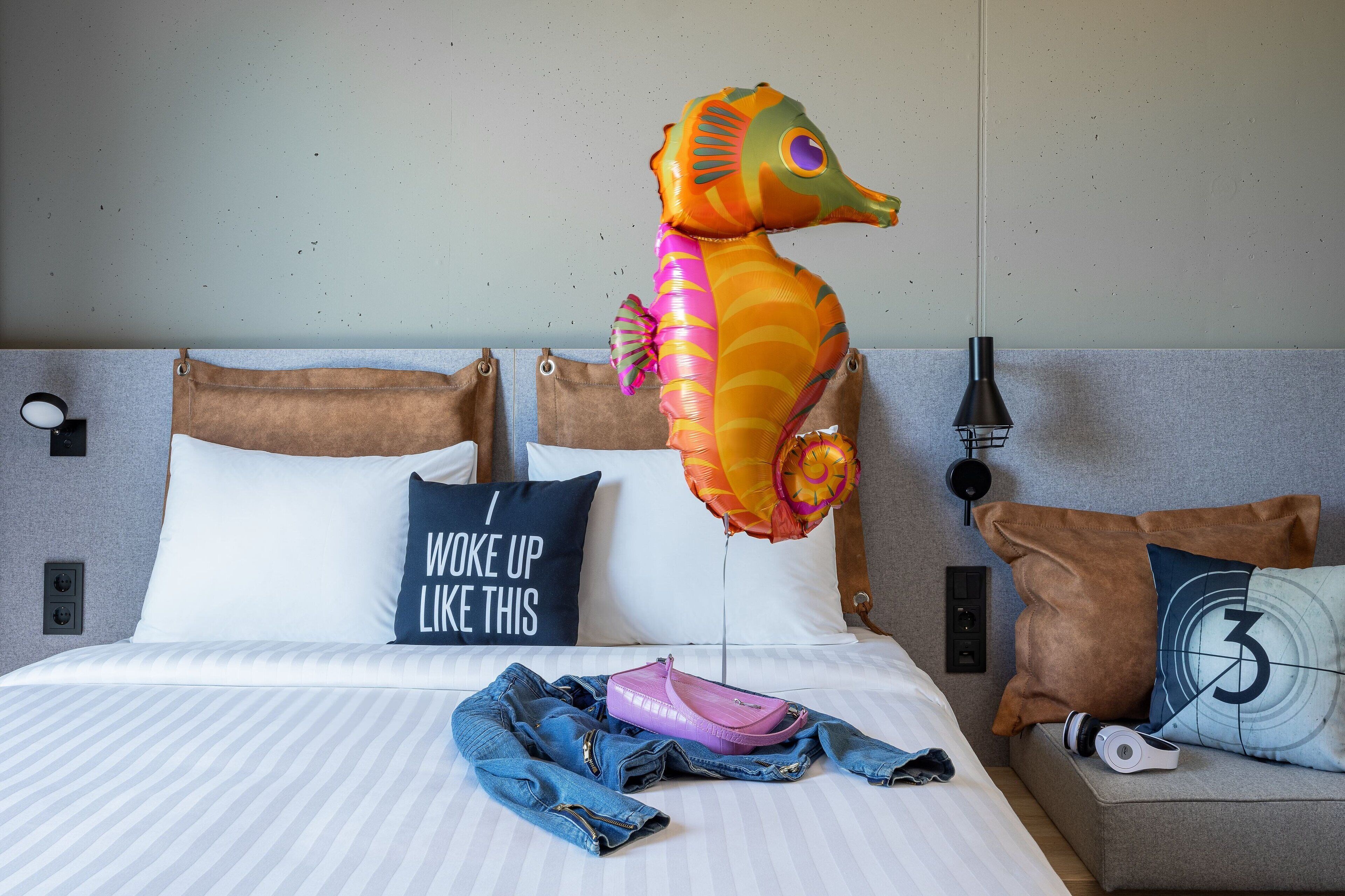 A bed with a striped bedspread, a pillow with the text "I woke up like this," and a seahorse balloon.