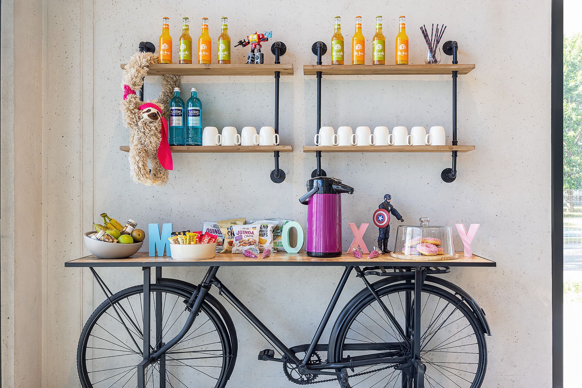 Una mesa tipo bicicleta con repisas que sostienen botellas, un cuenco de frutas y letras del alfabeto.
