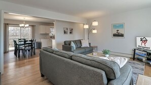 A modern living room with a grey sectional sofa, a dining table with chairs, a kitchen area, and a balcony with a view. - Pondside View Family Friendly Home!  [Moose Lodge] (Bar Harbor)