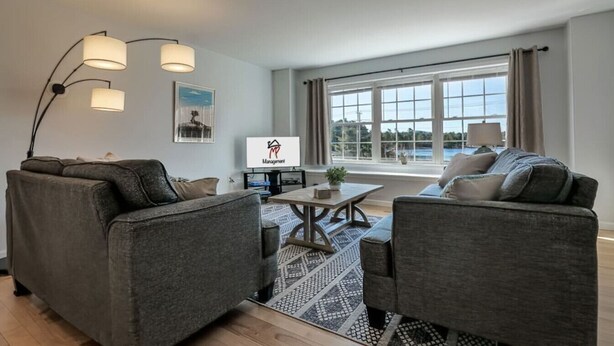A modern living room with two grey sofas, a coffee table, and a TV on a stand. There is a large window with curtains, a floor lamp, and a framed picture on the wall. - Pondside View Family Friendly Home!  [Moose Lodge] (Bar Harbor)