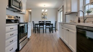 A modern kitchen with white cabinetry, a built-in oven, a microwave, and a dishwasher. There is a dining area with a table and chairs in the center. - Pondside View Family Friendly Home!  [Moose Lodge] (Bar Harbor)