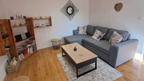 A spacious living room with a wooden floor, a grey sofa with patterned cushions, a coffee table, and a bookshelf.