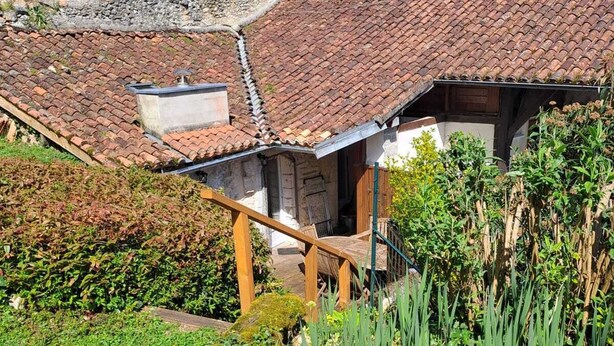 A rustic stone building with a tiled roof, a wooden staircase, and a garden with various plants.