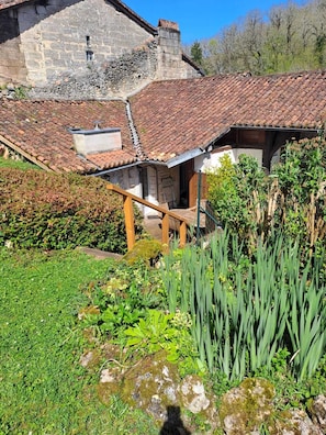 Un bâtiment rustique en pierre, doté d’un toit en tuiles, d’un escalier en bois et d’un jardin où poussent différentes espèces de plantes.