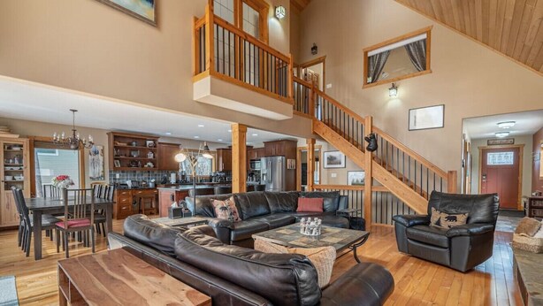 A spacious living area with a large black leather sofa, a wooden coffee table, and a dining table with chairs. A staircase leads to an upper level with a balcony. - 3 Bears Cabin | Games Room | Firepit | Lux Kitchen (Invermere)
