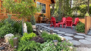 A wooden cabin with a stone patio, red Adirondack chairs, and a variety of plants. - 3 Bears Cabin | Games Room | Firepit | Lux Kitchen (Invermere)