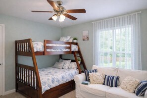 Una cama litera con ventilador de techo, un sofá blanco con almohadas a rayas y una ventana con cortinas traslúcidas.