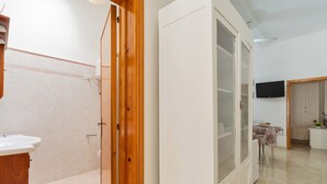 A room with a wooden door, a white cabinet, a tiled floor, a television mounted on the wall, and a table with a tablecloth.