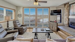 A living room with a fireplace, a ceiling fan, and a large window with blinds.
