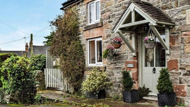 A stone house with a white door, a small porch, and a window with a basket of flowers. - Cosy & Spacious Cottage in Scenic Village With Pub (Ashbourne)