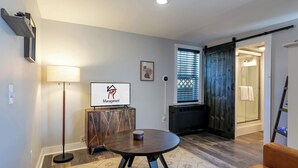 Una sala de estar moderna con una mesa redonda, una lámpara de pie, un televisor mostrando un logotipo, una ventana con persianas, una puerta corrediza y una zona de ducha.