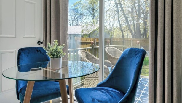 Una habitación con una mesa de vidrio y dos sillas azules, una planta en maceta, y vista a un jardín trasero con mobiliario de mimbre.