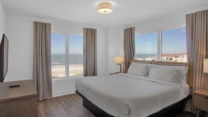 Ein Hotelzimmer mit einem großen Bett, einem Fernseher, einem Nachttisch und Blick auf den Strand.