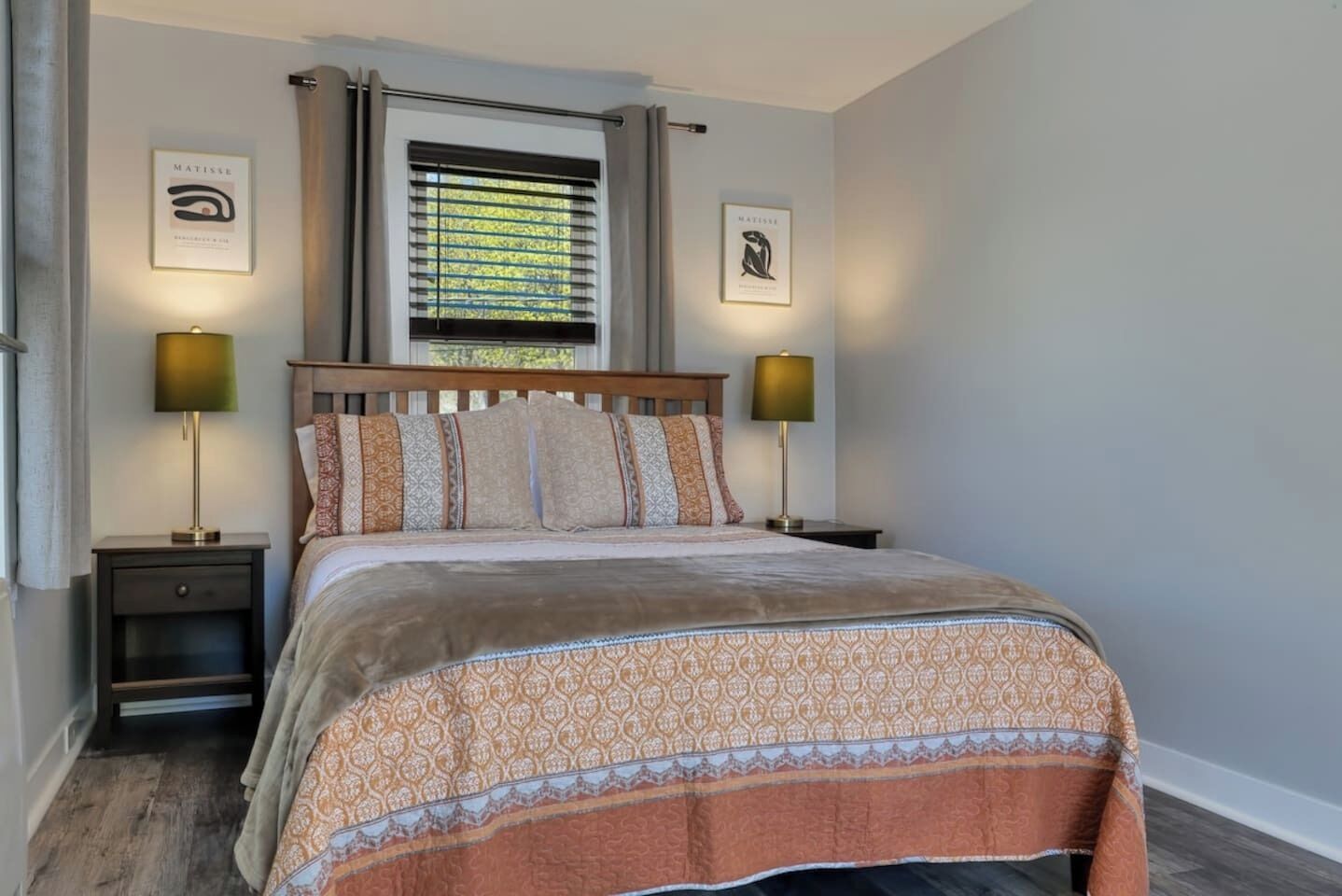 A bedroom with a bed, two bedside tables, a window with blinds, and two wall-mounted lamps.