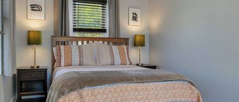 A bedroom with a bed, two bedside tables, a window with blinds, and two wall-mounted lamps.