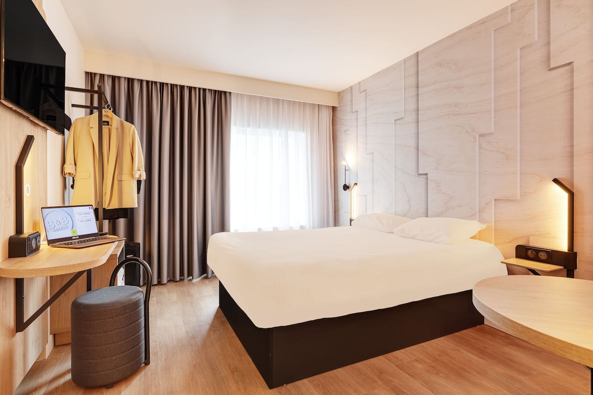 Moderni hotellihuone, jossa on suuri sänky, työpöytä kannettavalla tietokoneella, tuoli ja seinälle kiinnitetty naulakko.