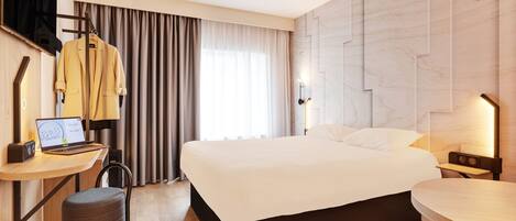 Une chambre d’hôtel moderne avec un grand lit, un bureau avec un ordinateur portable, une chaise et un porte-manteau mural.