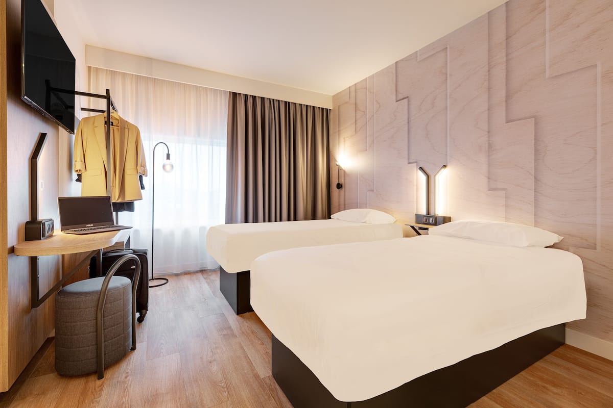 Moderni hotellihuone, jossa on kaksi sänkyä, työpöytä kannettavalla tietokoneella, tuoli ja seinälle asennettu televisio.