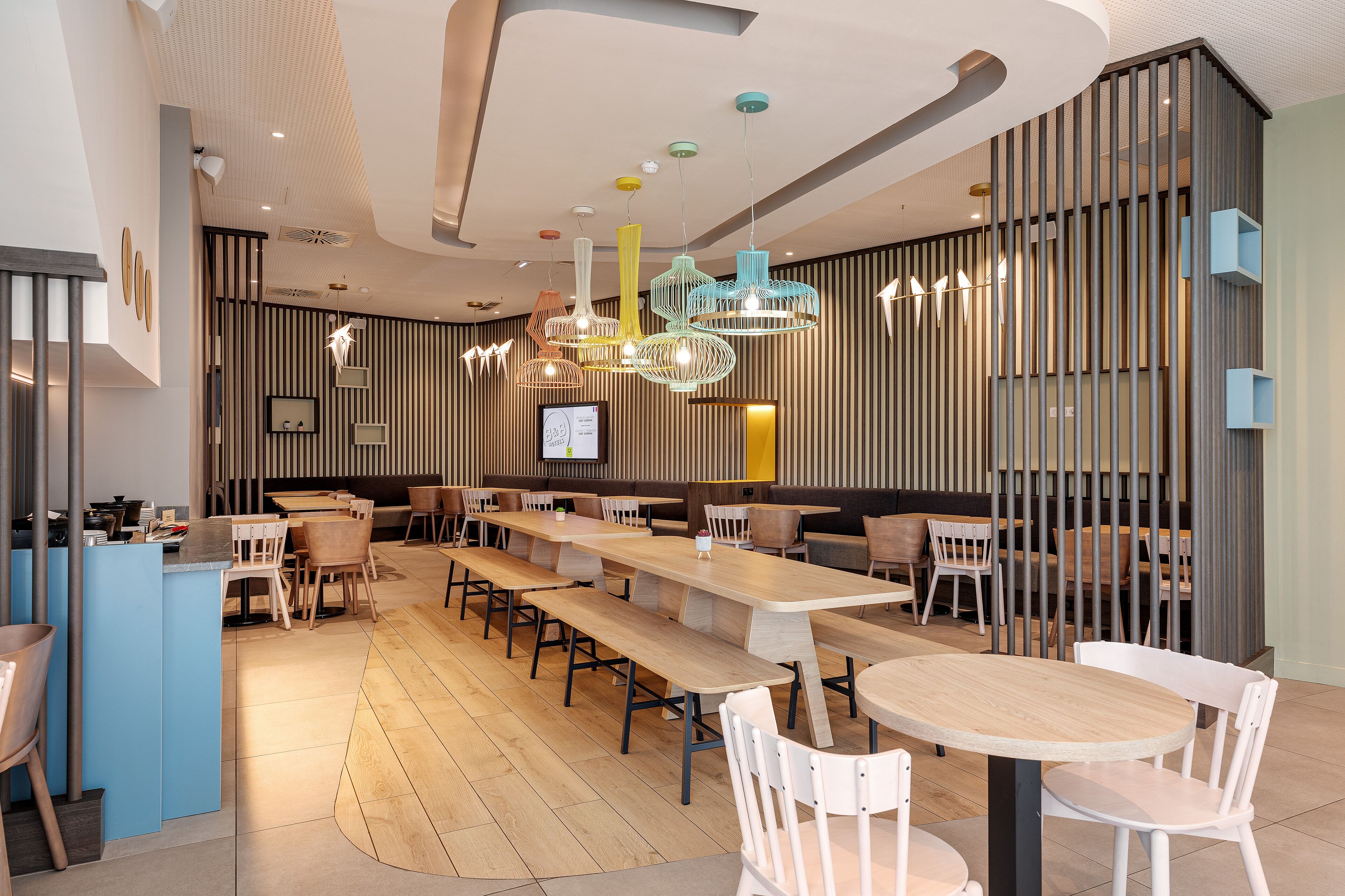 A modern restaurant with long communal tables, high ceilings with unique lighting fixtures, and a mix of seating options including booths and chairs.