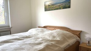A bedroom with a wooden bed, a white textured blanket, a nightstand with a lamp, and a window with a view of a landscape. - Apartment "Alpenglöckchen", Oberstaufen/Steibis (Oberstaufen)