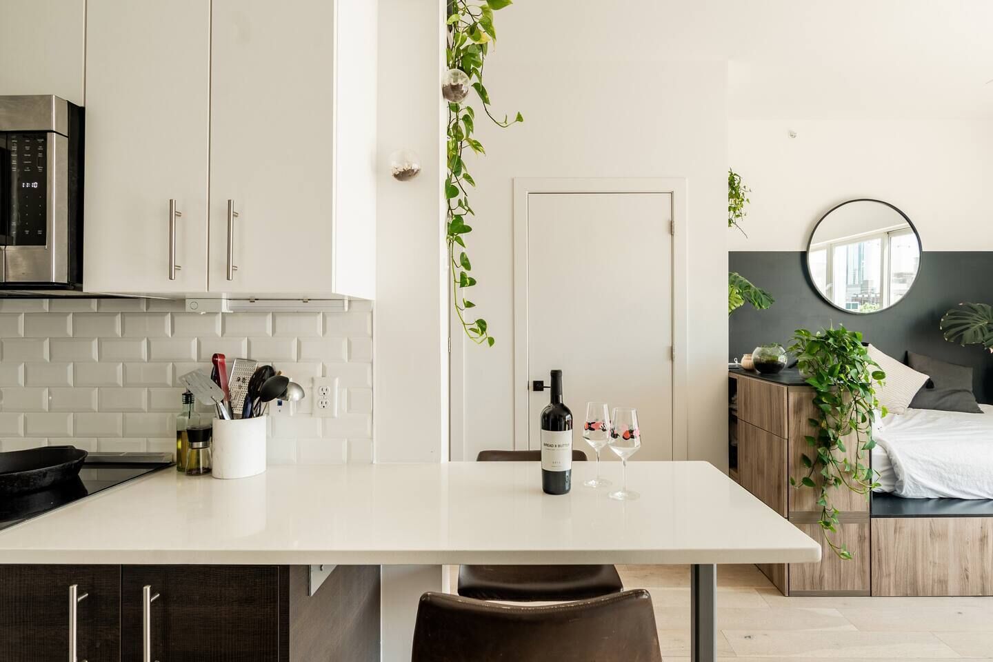 Ett modernt kök med vita skåp, en rostfri köksfläkt och en vit bänkskiva. Det finns ett matbord med en vinflaska och två glas. Rummet har en minimalistisk design med en grå vägg och en rund spegel.
