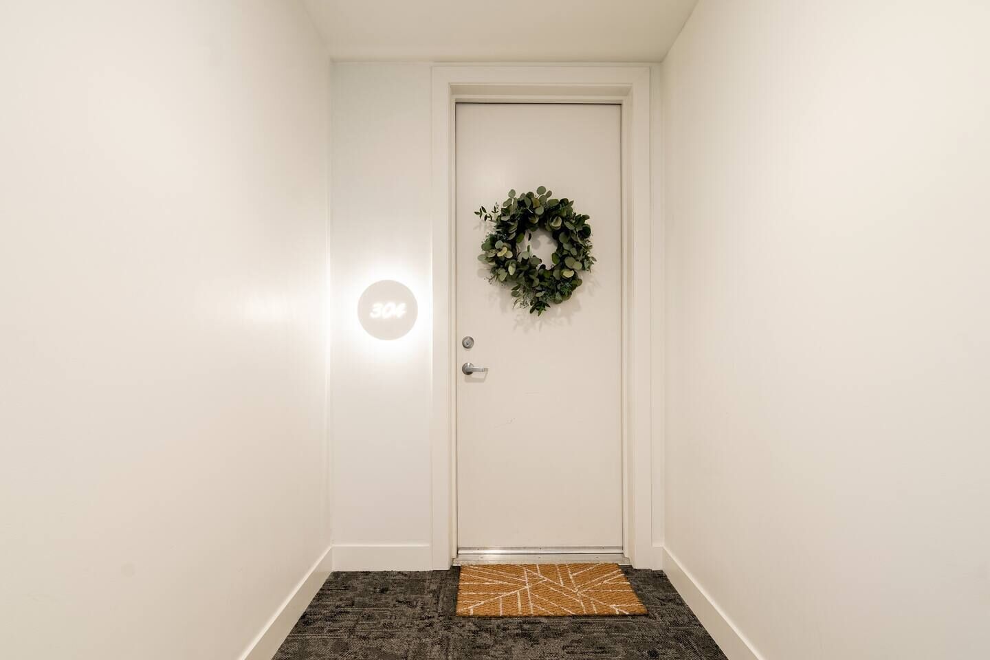A white door with a number plate, a wreath, and a textured doormat.