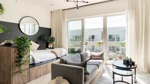 Ein modernes Schlafzimmer mit einem Bett, einem grauen Sofa und einem kleinen Tisch. An den Wänden befinden sich Pflanzen und ein Spiegel.