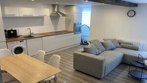 Een moderne keuken met witte kasten, een houten aanrecht en een ingebouwde oven. Er is een grijze bank met kussens, een kleine ronde tafel en een wasmachine.