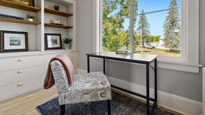 A study room with a desk, chair, shelving, and a large window with a view of trees and a house.