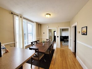 Un comedor con una mesa de madera larga y sillas, una ventana con cortinas y un pasillo que conduce a una sala de estar.