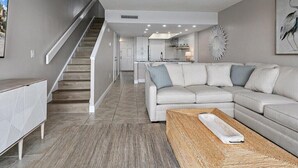 A modern living room with a staircase, a sectional sofa, a coffee table, and a kitchen in the background. - Island House A209 (St. Augustine)