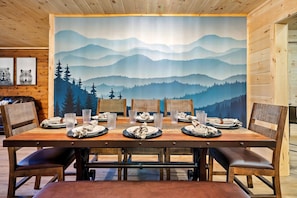 A dining table set with plates, glasses, and cutlery, surrounded by chairs, with a scenic mountain view mural on the wall.