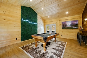 A pool table in a wooden room with a green wall and a painting of mountains.