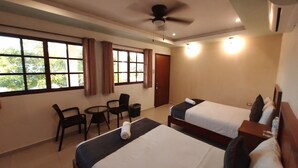 Habitación de hotel con dos camas, una mesita con sillas, un ventilador de techo y ventanas amplias con cortinas.