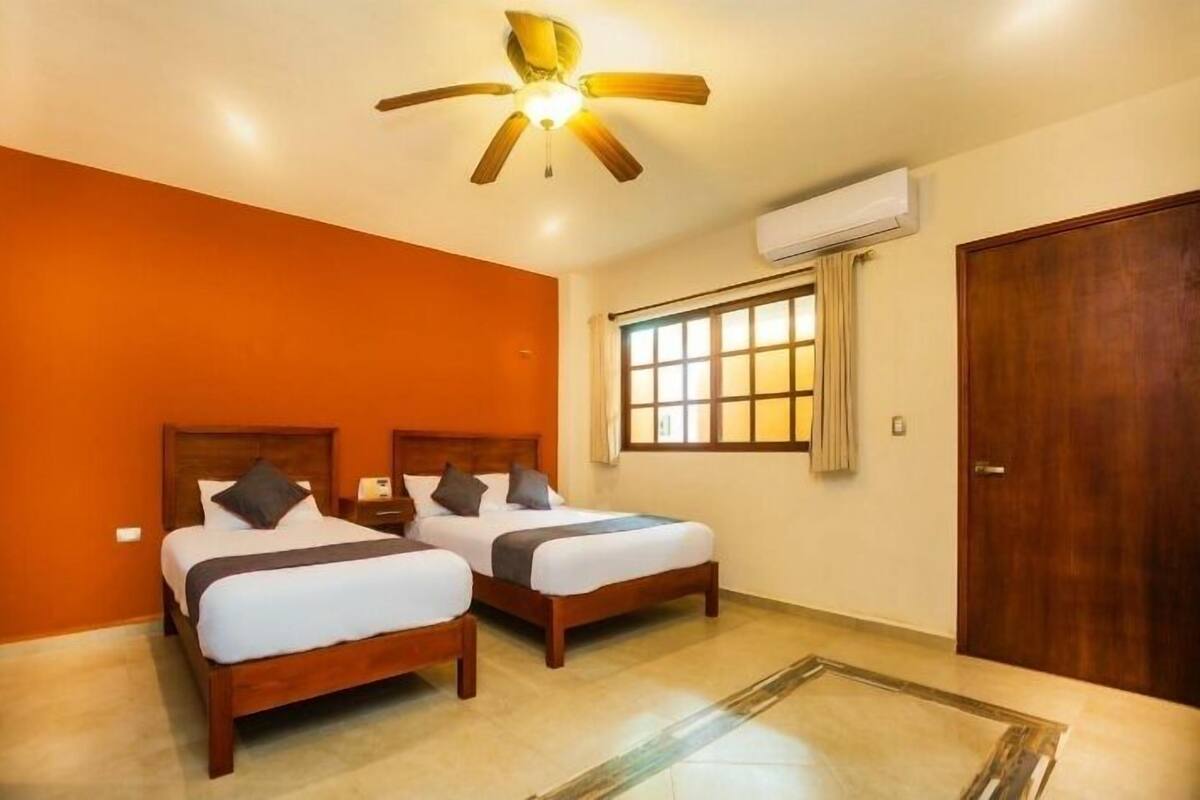 Una habitació d'hotel amb dos llits de fusta, una paret d'accent taronja, un ventilador de sostre i una finestra amb cortines.