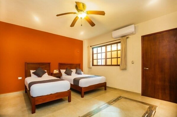 Habitación de hotel con dos camas de madera, una pared de acento naranja, un ventilador de techo y una ventana con cortinas.