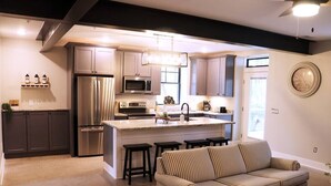 A modern kitchen with stainless steel appliances, a central island, and a striped sofa in a living area. - The Hemlock Loft (Boyne City)