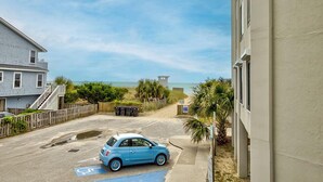 A parking area with a blue car, a building with a beige exterior, and a view of the beach with palm trees. - Ocean Views, Balcony, Pool, STEPS to Beach, Restaurants, Shops (Wrightsville Beach)