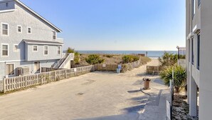 A two-story building with a flat roof and a balcony, adjacent to a sandy area with a fence and a trash bin. - Ocean Views, Balcony, Pool, STEPS to Beach, Restaurants, Shops (Wrightsville Beach)